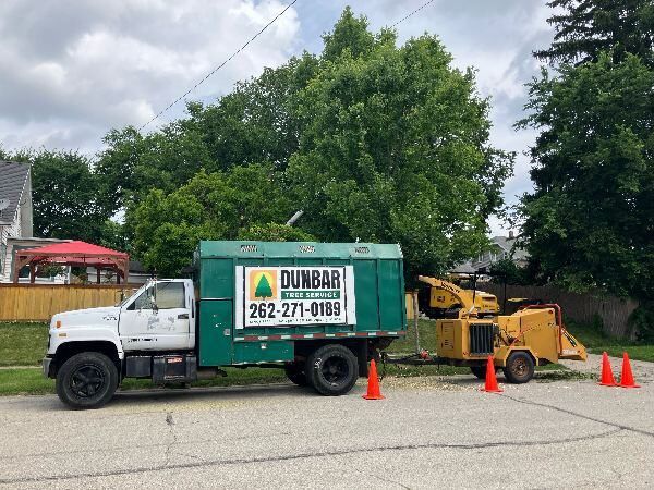 Dunbar Tree Service truck with logo and contact number parked on a residential street, accompanied by a wood chipper and surrounded by safety cones, emphasizing professional tree care equipment in Waukesha.